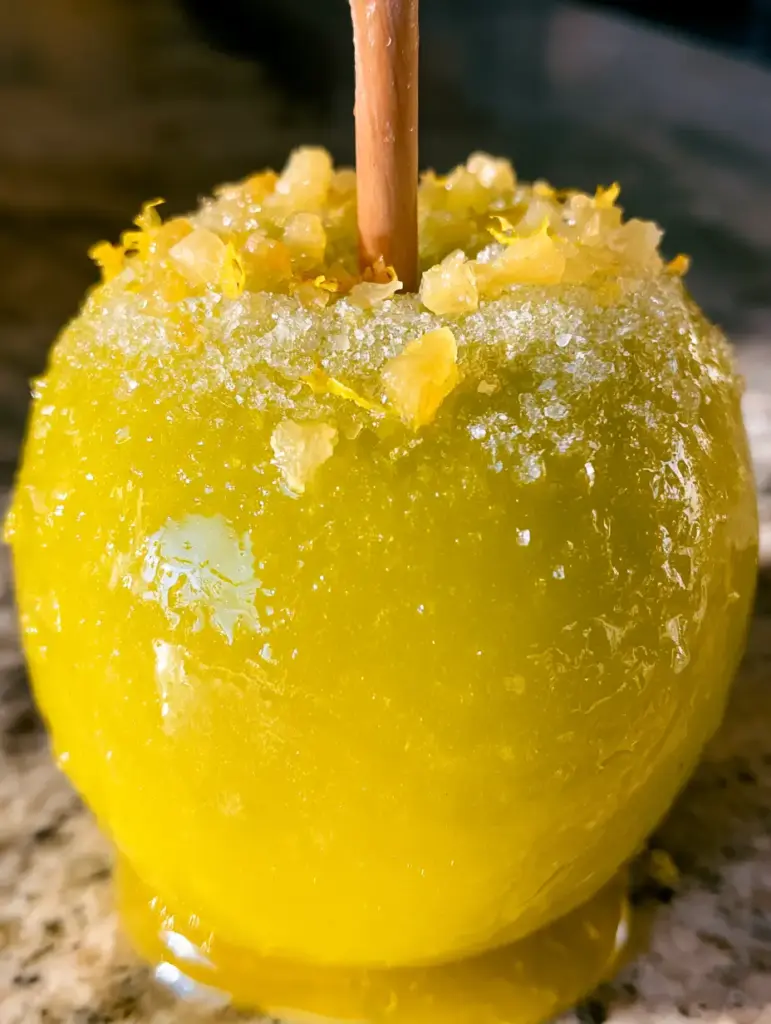 Pineapple Lemonade Glazed Apples: The Ultimate Neon Summer Candy Apple 1 A single pineapple lemonade glazed apple on a kitchen counter showing the translucent yellow coating and wooden stick.