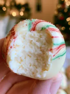 A close-up handheld shot of a white chocolate cheesecake truffle with a bite taken out, showing the dense white filling and holiday sprinkles. (cheesecake truffles)
