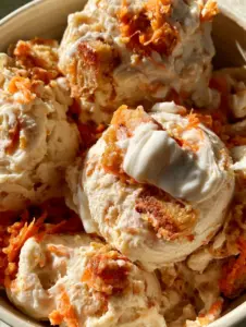 A handheld smartphone photo of a luxurious bowl of homemade carrot cake ice cream with golden cake bits and a thick frosting swirl.