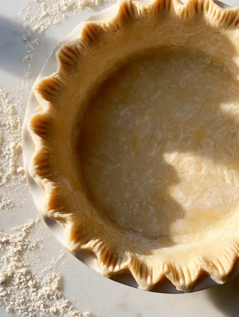 Pie Crust Recipe: The Ultimate Guide to Buttery Flaky Pastry 1 Top down shot of a raw pie crust in a white ceramic dish on a white marble surface.